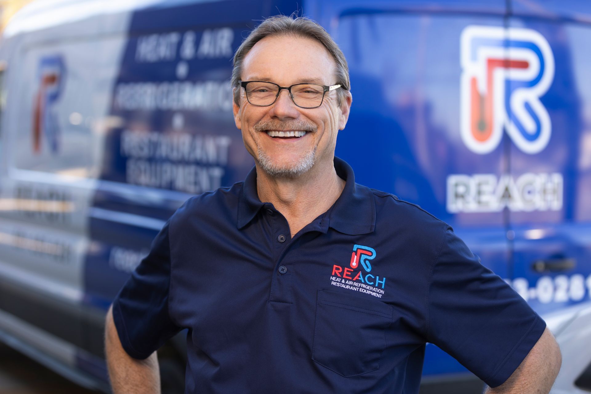 A smiling person in a dark blue REACH polo shirt stands in front of a blue REACH company van.