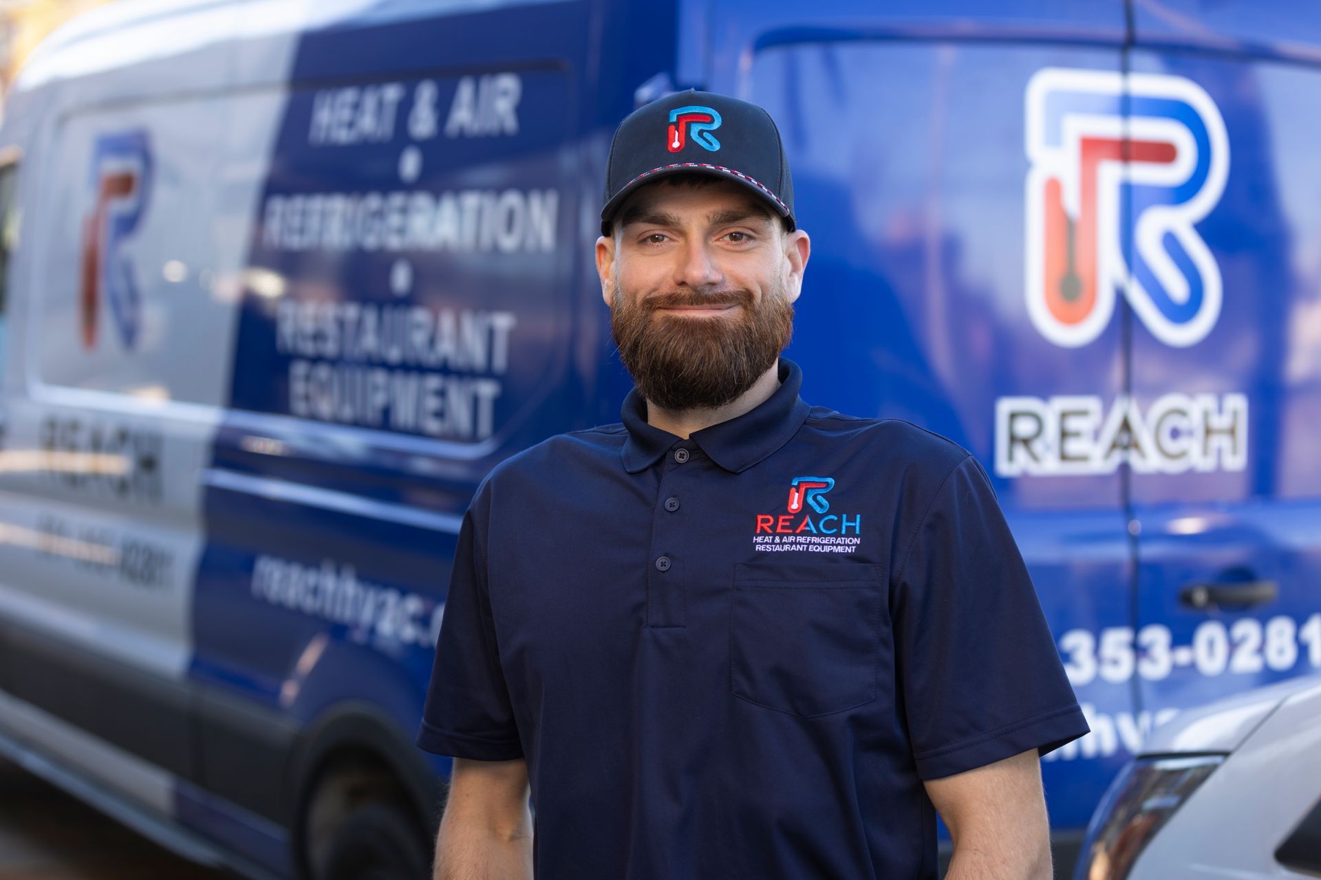 A technician in a branded navy polo and cap stands in front of a Reach Heat, Air & Refrigeration service van.
