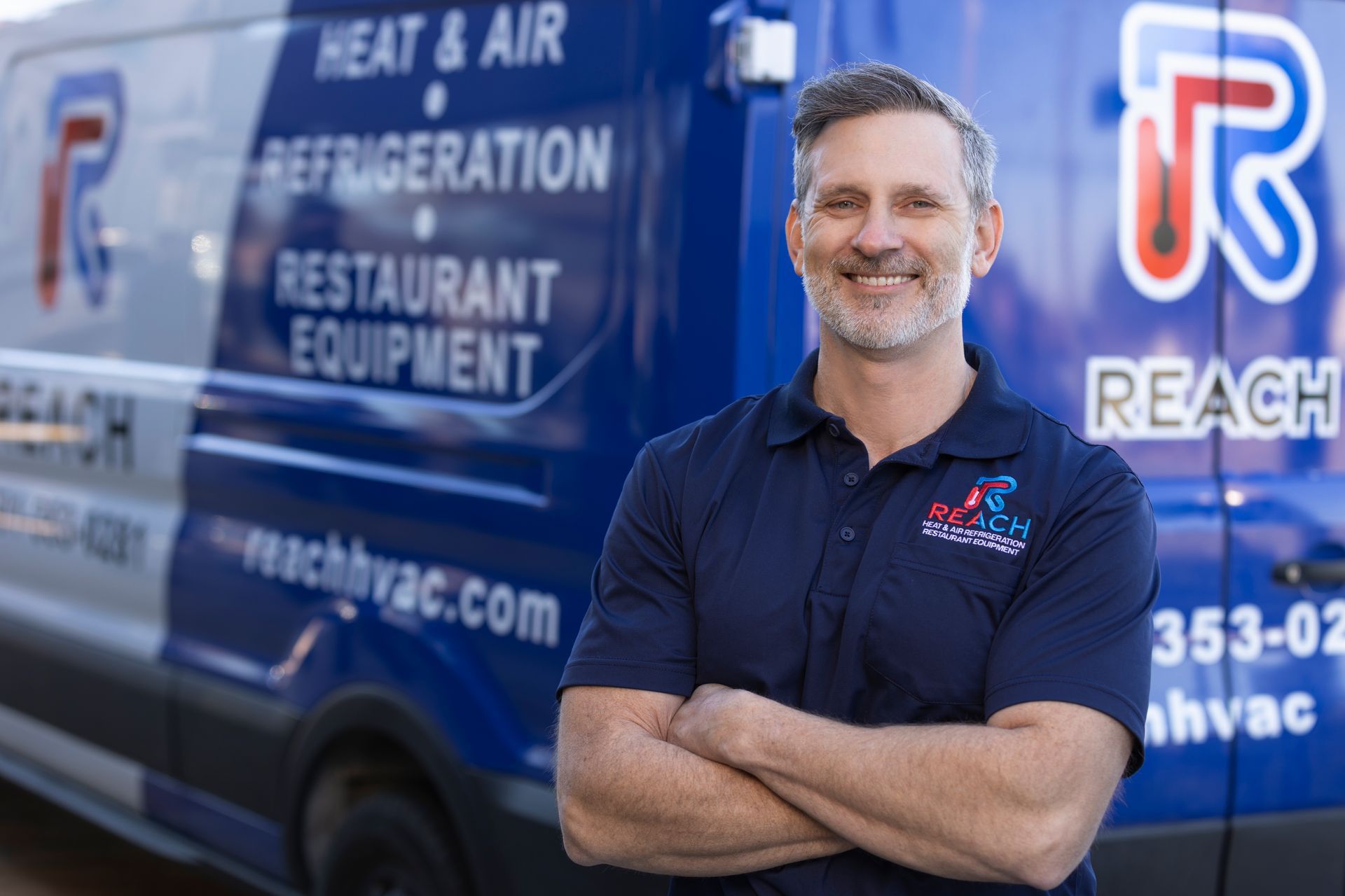 A smiling professional with arms crossed in front of a blue Reach Heat & Air service van.