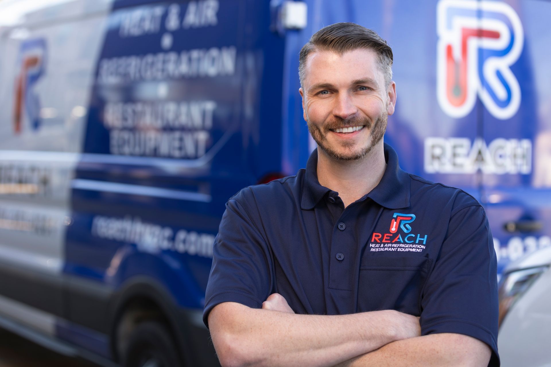 A smiling technician in a navy blue branded polo shirt stands with arms crossed in front of a company van.