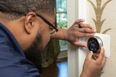 A person wearing glasses adjusts a round, digital smart thermostat mounted on a patterned wall.