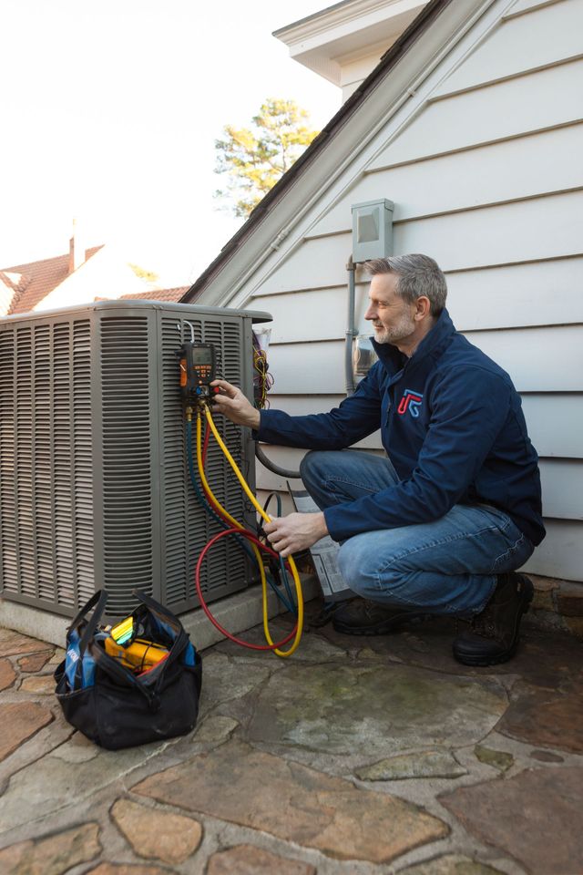 A technician in a blue jacket inspects an outdoor HVAC unit using a digital manifold gauge with yellow and red hoses.