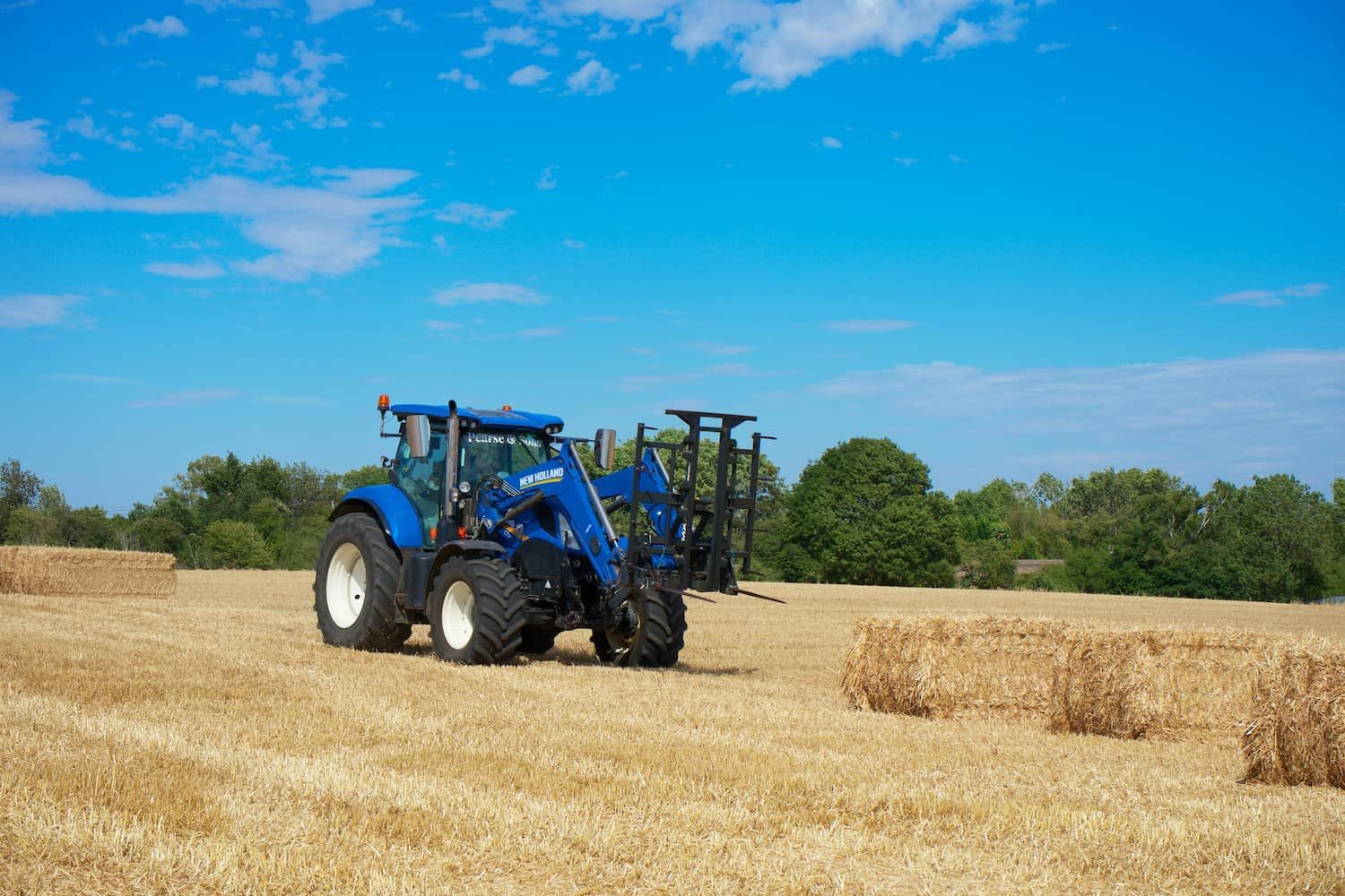 A Blue Tractor Is Driving Through a Field of Hay Bales — Parkes Machinery Services PTY LTD in Orange, NSW