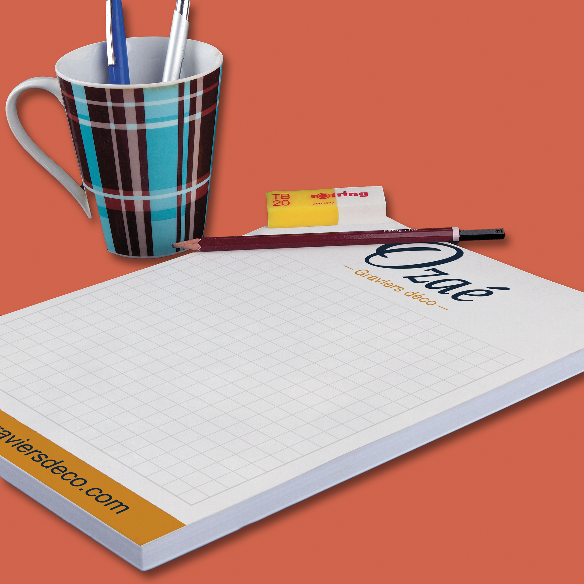 Bloc-notes quadrillé, tasse avec stylos et crayon sur fond orange. Le bloc-notes est orné du logo en haut et en bas.