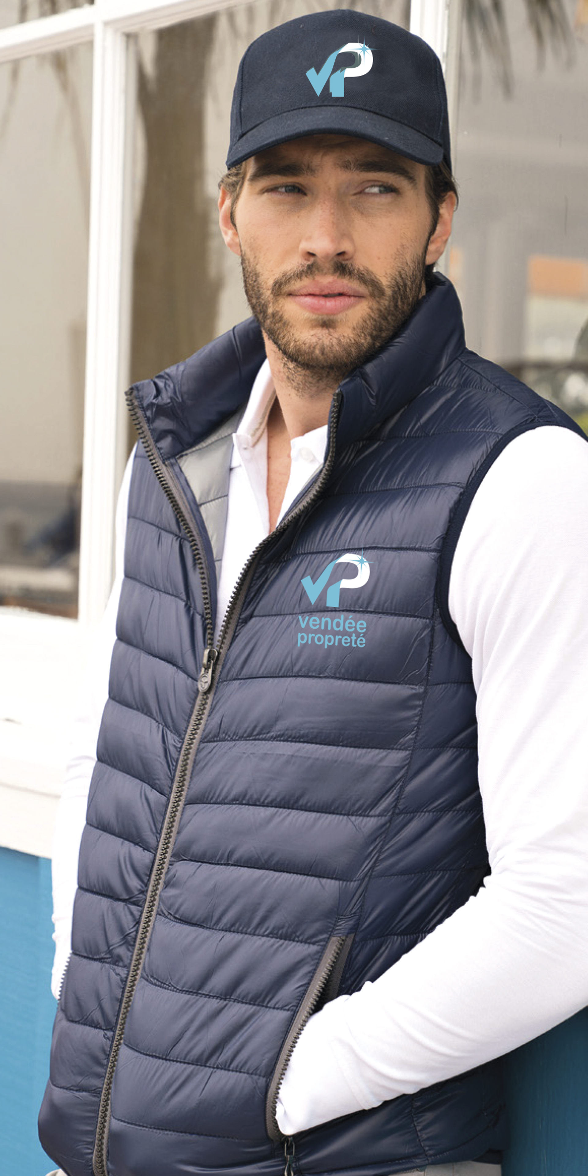 Un homme portant un gilet bleu marine et une casquette avec un logo, debout à l'extérieur. Il porte une chemise blanche.