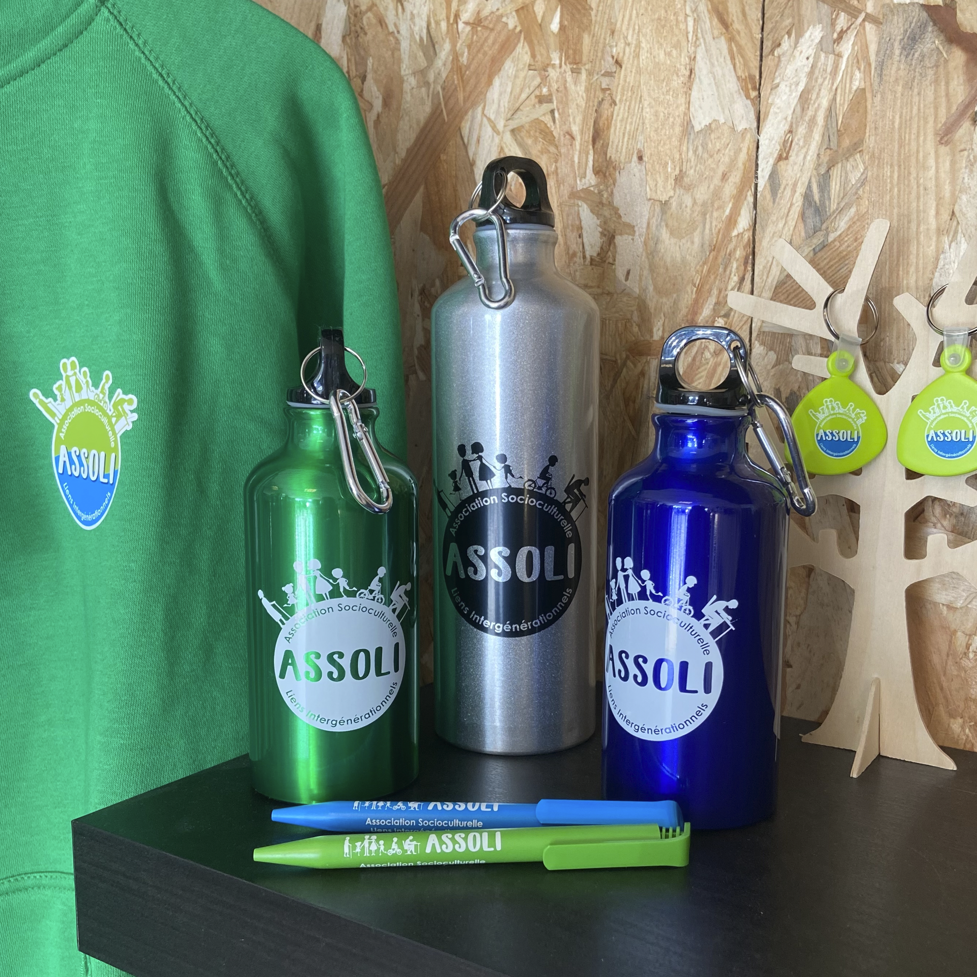 Sweat-shirt vert, bouteilles d'eau vertes, argentées et bleues, stylos et porte-clés avec le logo « ASSOLI » sur une étagère.