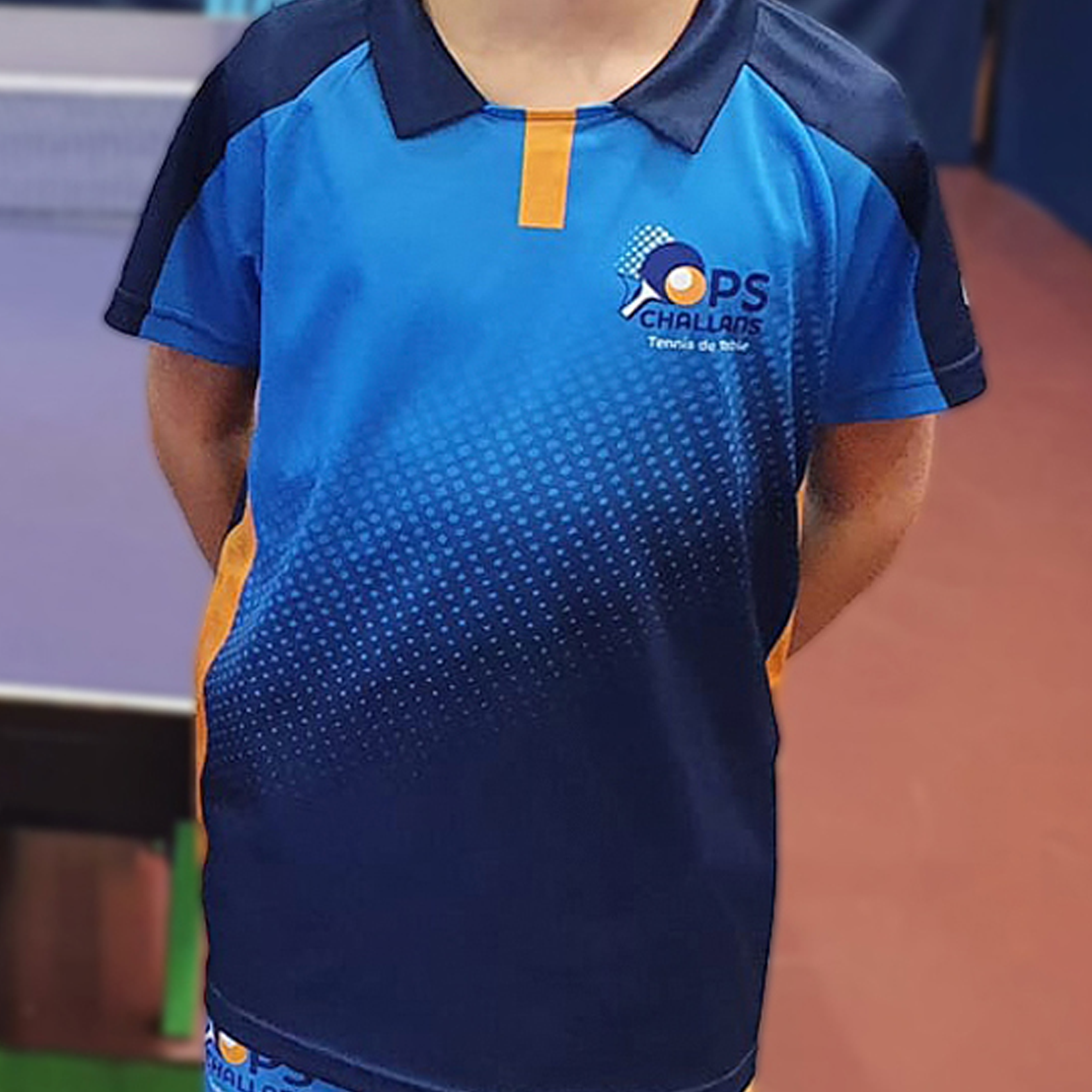 Un enfant portant une chemise de tennis de table bleue et bleu marine avec un logo, debout dans un décor de tennis de table.