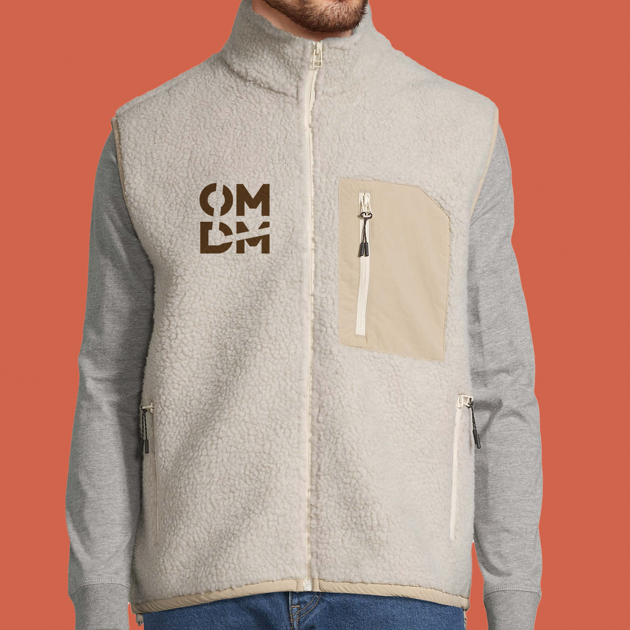 Homme portant un gilet polaire beige, une chemise grise à manches longues et un jean. Le gilet a un logo et d'une poche.