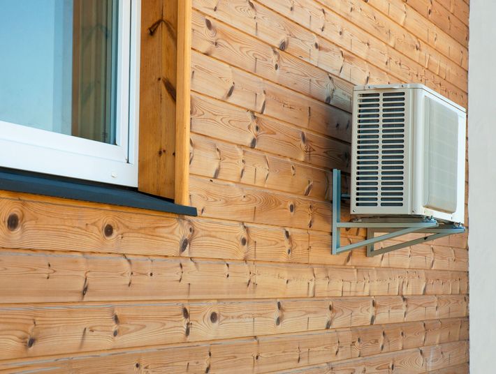 Outdoor mini-split AC condenser unit mounted on wood siding wall beside window.