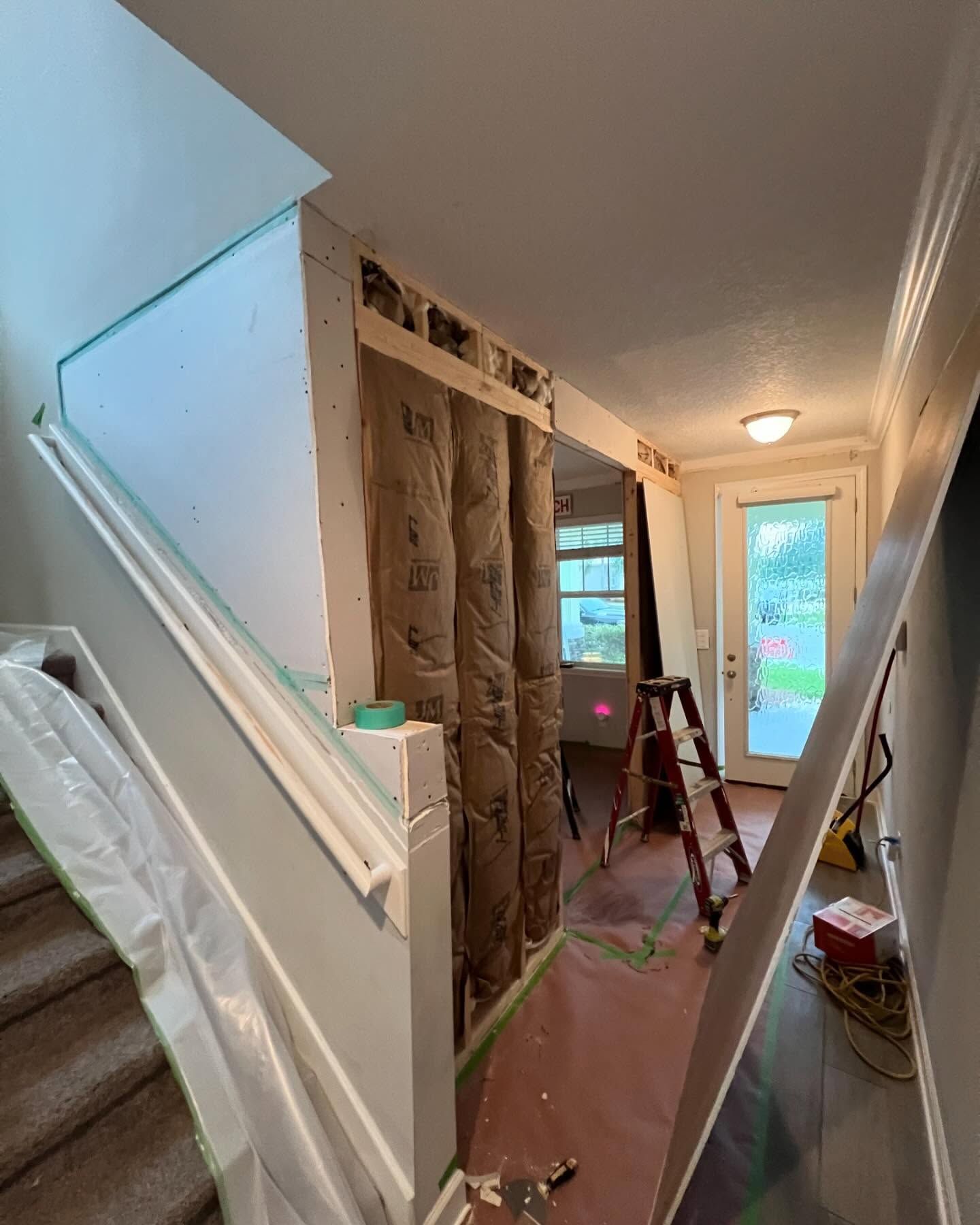 Hallway under construction: Stairway to the left, open wall with insulation, door and ladder visible in the distance.