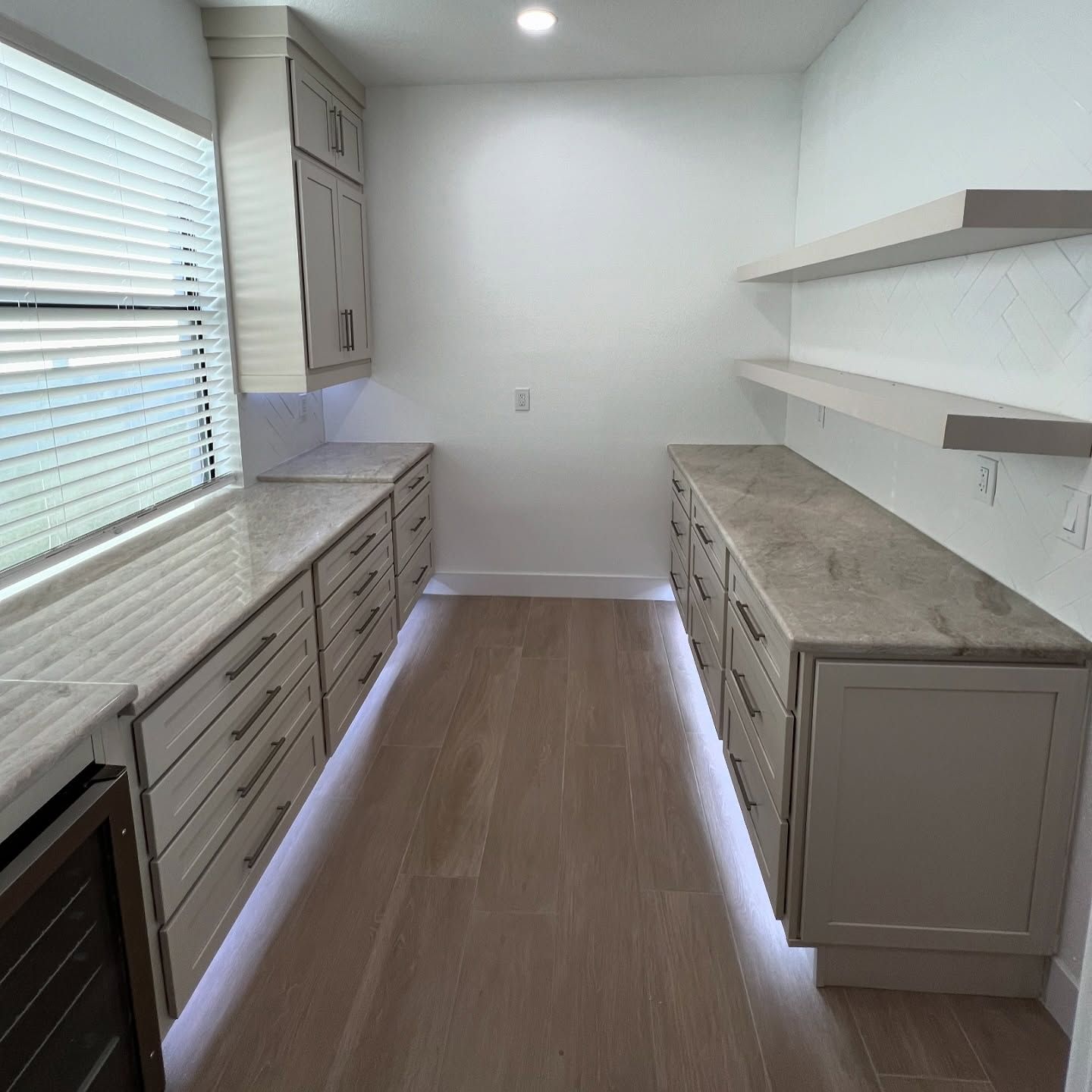 Pantry with light gray cabinets, drawers, and countertops. Floating shelves and a window. Wooden floors.