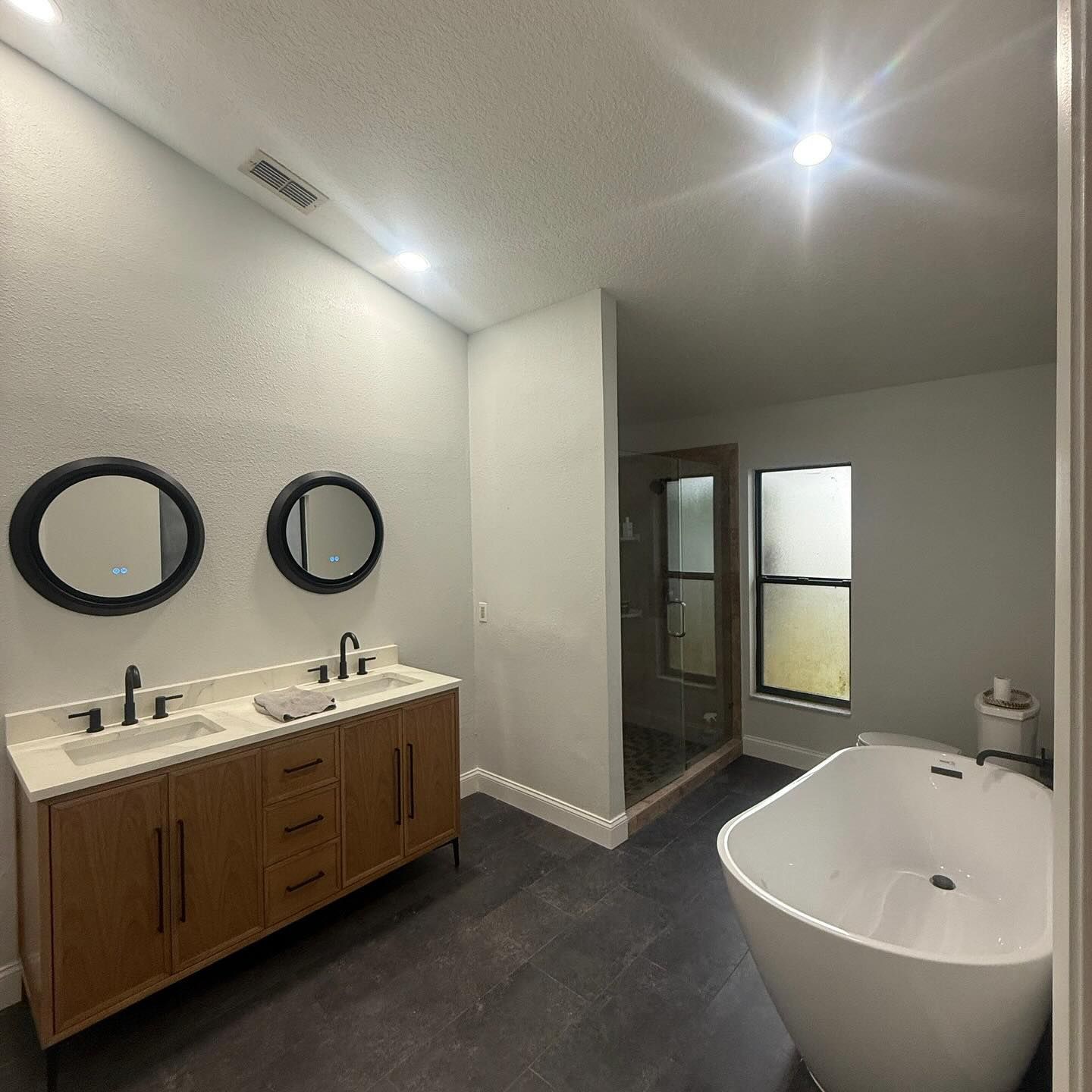 Modern bathroom with wooden double vanity, two round mirrors, freestanding tub, and shower.