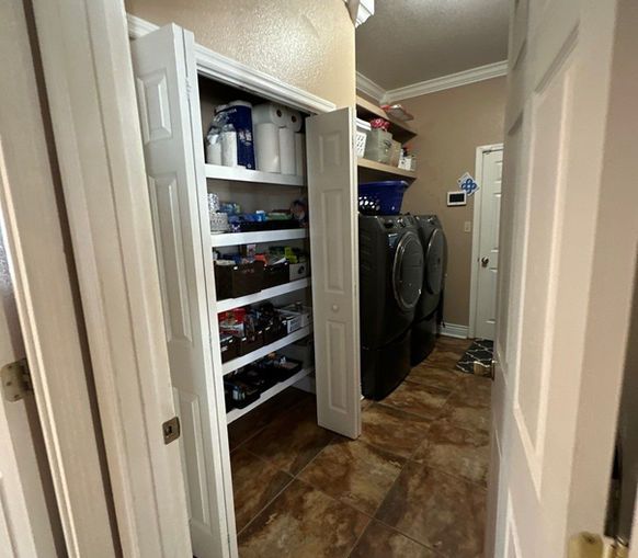 Laundry room with open pantry door, washer/dryer, and shelves of supplies. Brown tile floors.