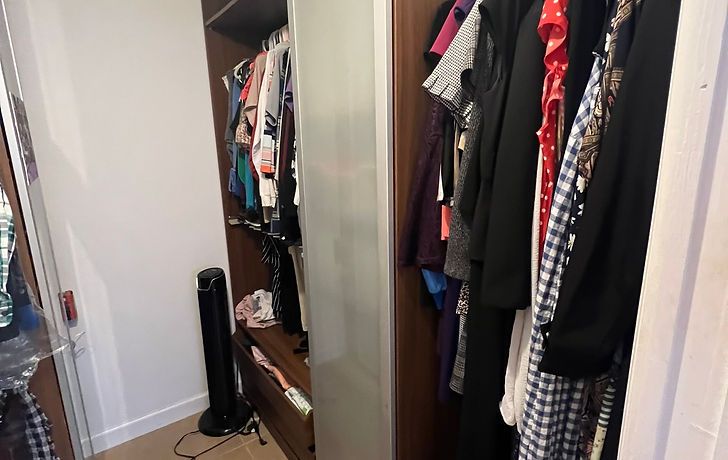 Closet with hanging clothes of various colors and a black fan on the floor.