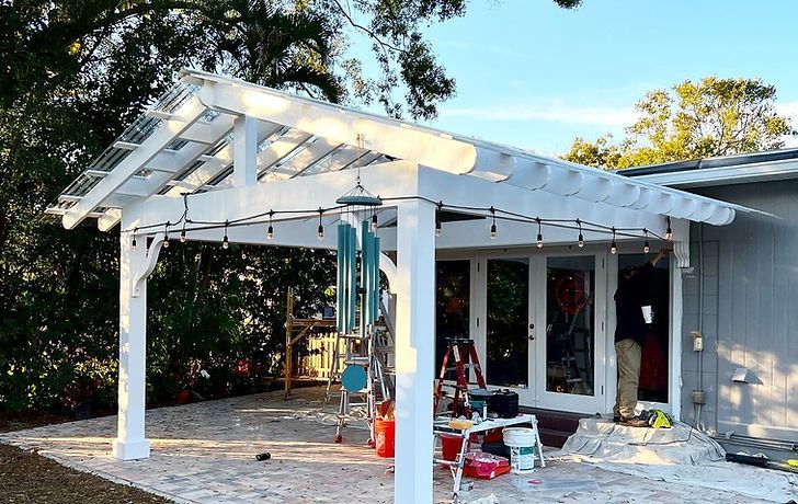 White pergola with transparent roof over a patio, strung with lights.  A person works inside a nearby doorway.