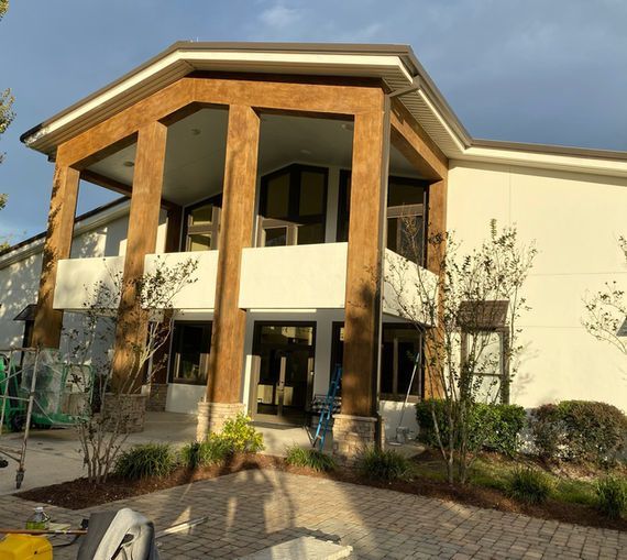 Two-story building with white stucco walls, brown columns, and large windows; daytime, outdoor setting.