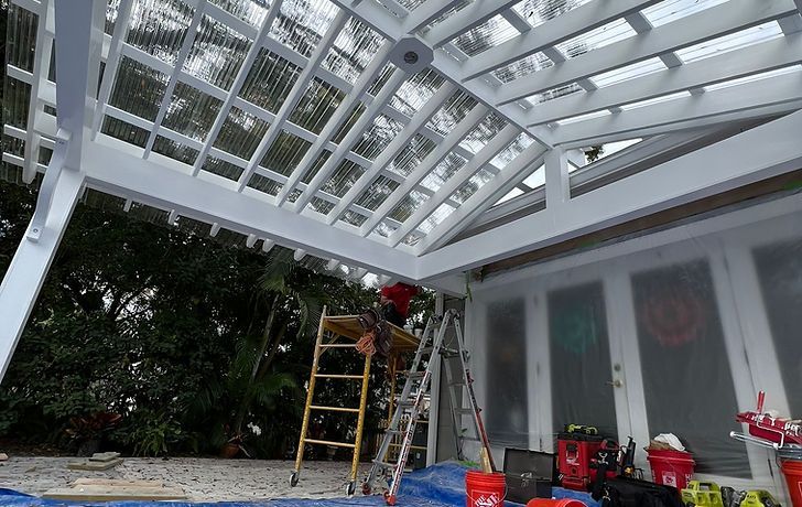 Person on a ladder painting a white pergola with a clear, corrugated roof. Outdoor setting.
