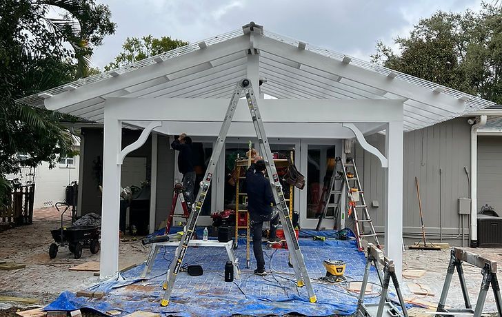 Construction workers installing a white pergola over a patio. Tools, ladders, and tarps are visible.