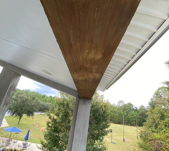 Angled view of a building's underside with a wood-toned beam and white ceiling. Green trees and a blue umbrella are in the background.