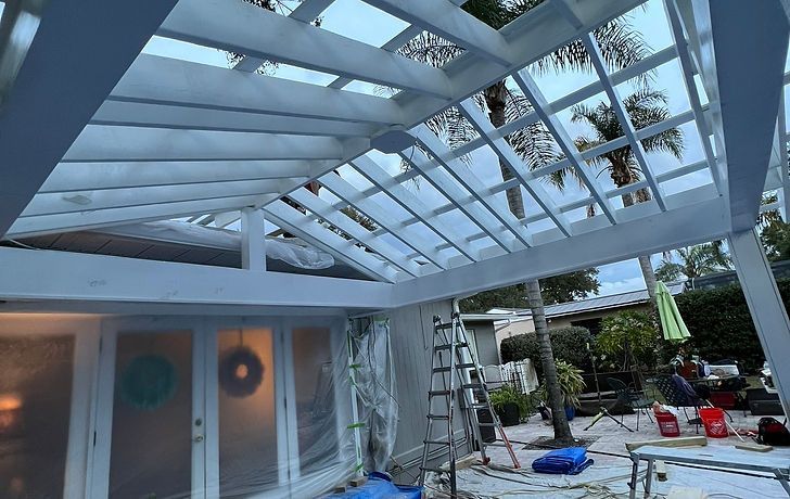 White painted pergola over patio with glass doors, ladder, and trees in background.