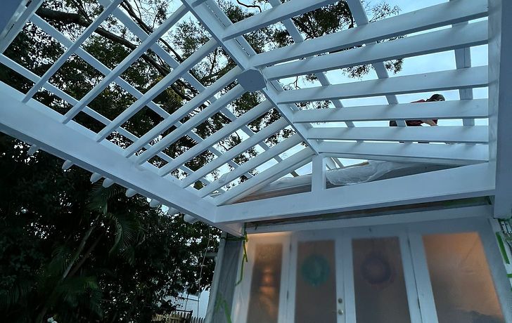 White pergola over a building with white doors and frosted windows. Green trees in the background.