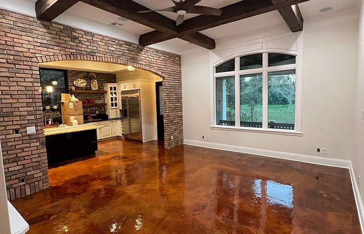 Room with brick archway, glossy brown floor, white trim, exposed wooden beams, and a large window overlooking greenery.