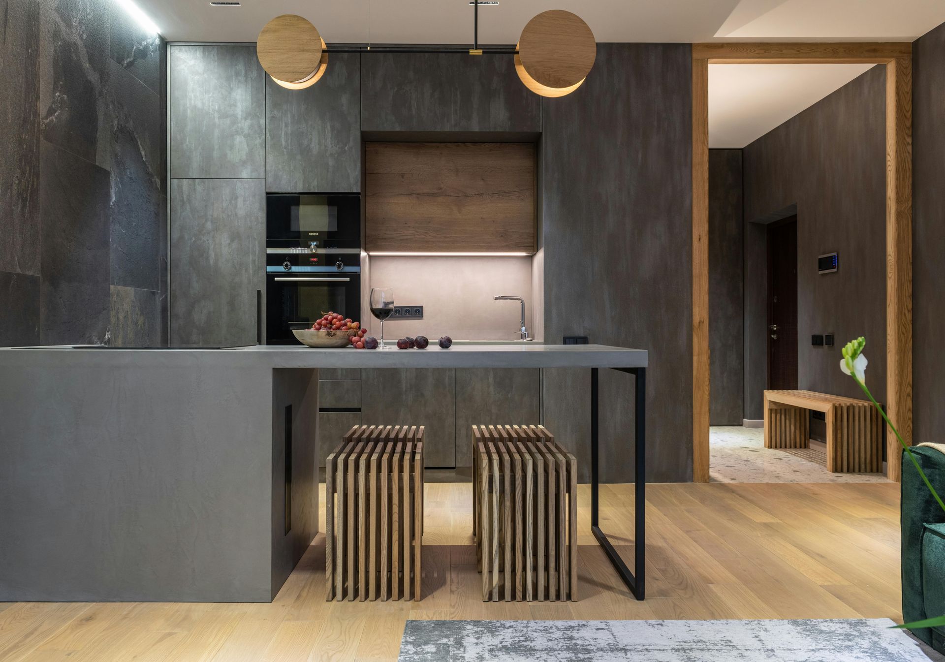 Modern kitchen with gray stone walls, wood accents, island with two stools, and a glimpse of a hallway.