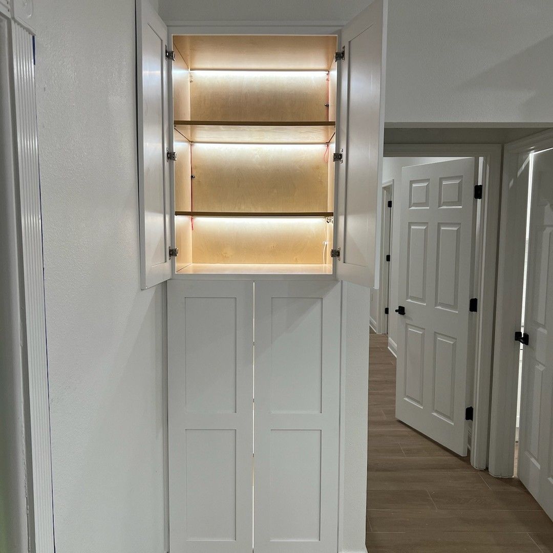 White cabinet with open upper doors and interior lighting, two glass shelves. Lower cabinet doors are closed. Hallway in the background.