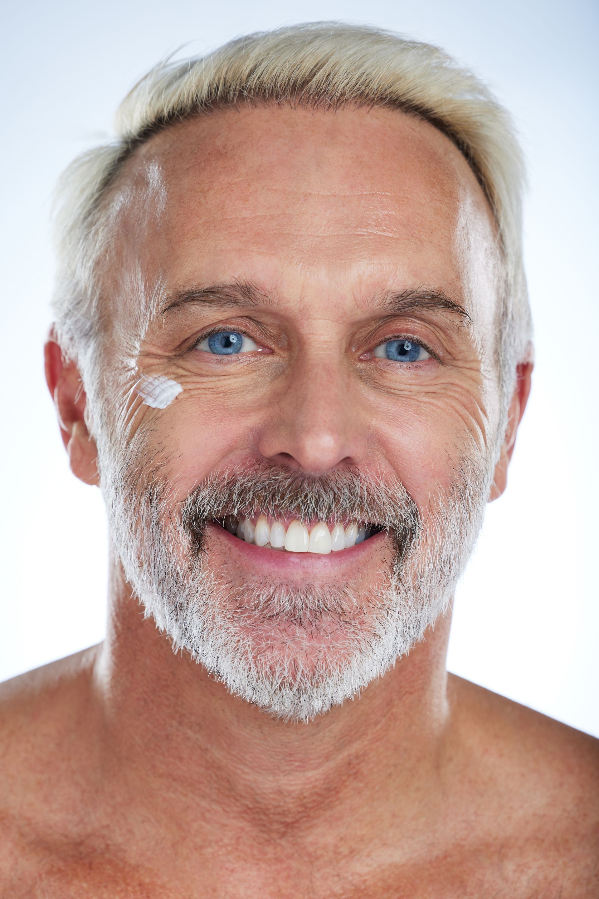 a shirtless man with a beard is smiling and applying lotion to his face .