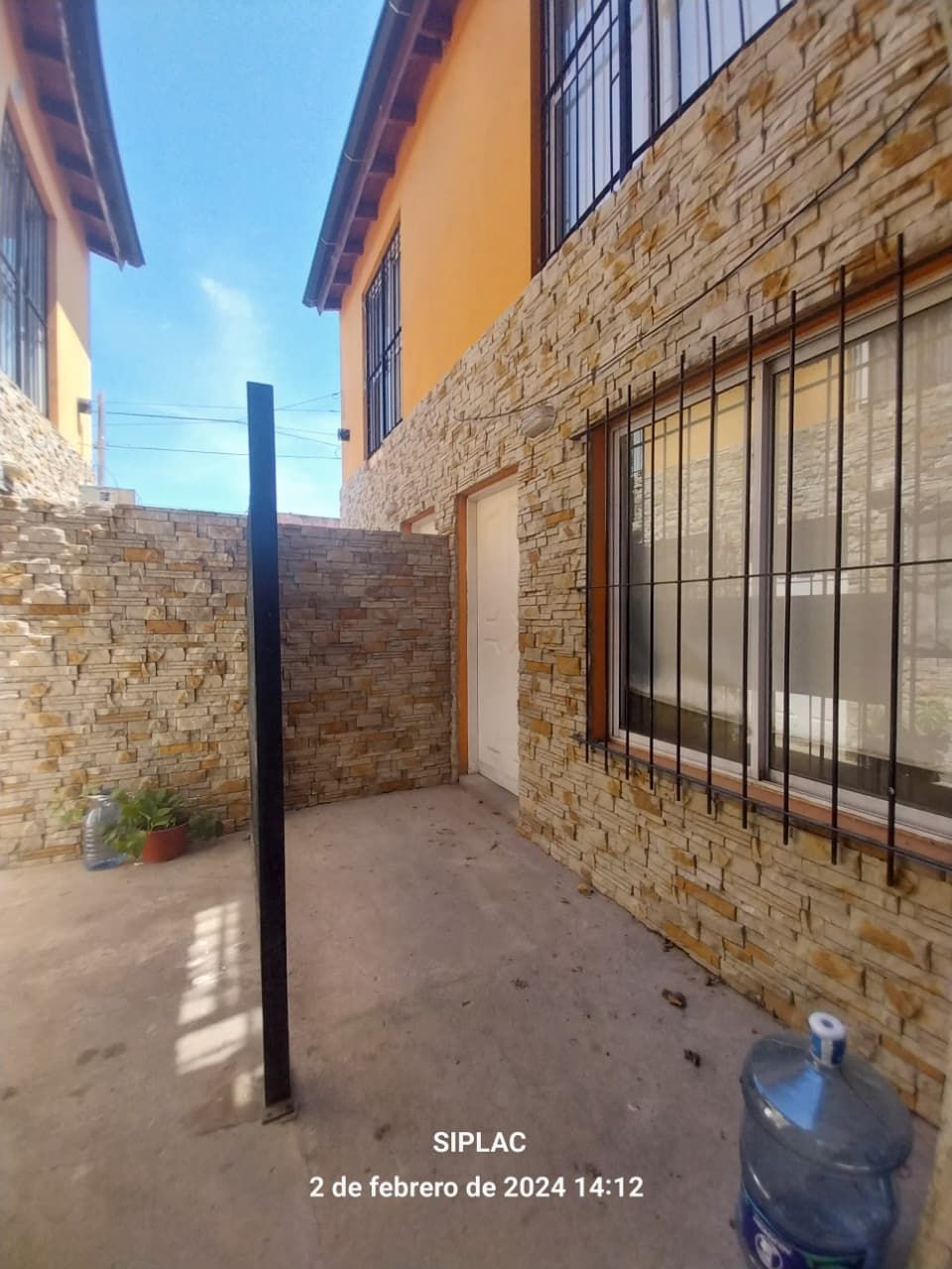 Exterior de edificios de dos pisos con paredes de piedra y color naranja, rejas de metal y una jarra de agua en el suelo.