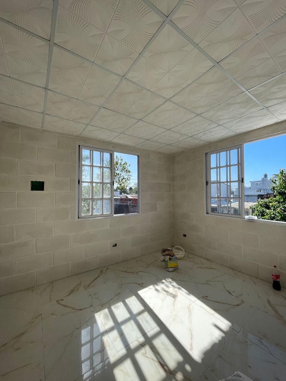 Habitación vacía con dos ventanas y suelo de baldosas; construcción sin terminar.