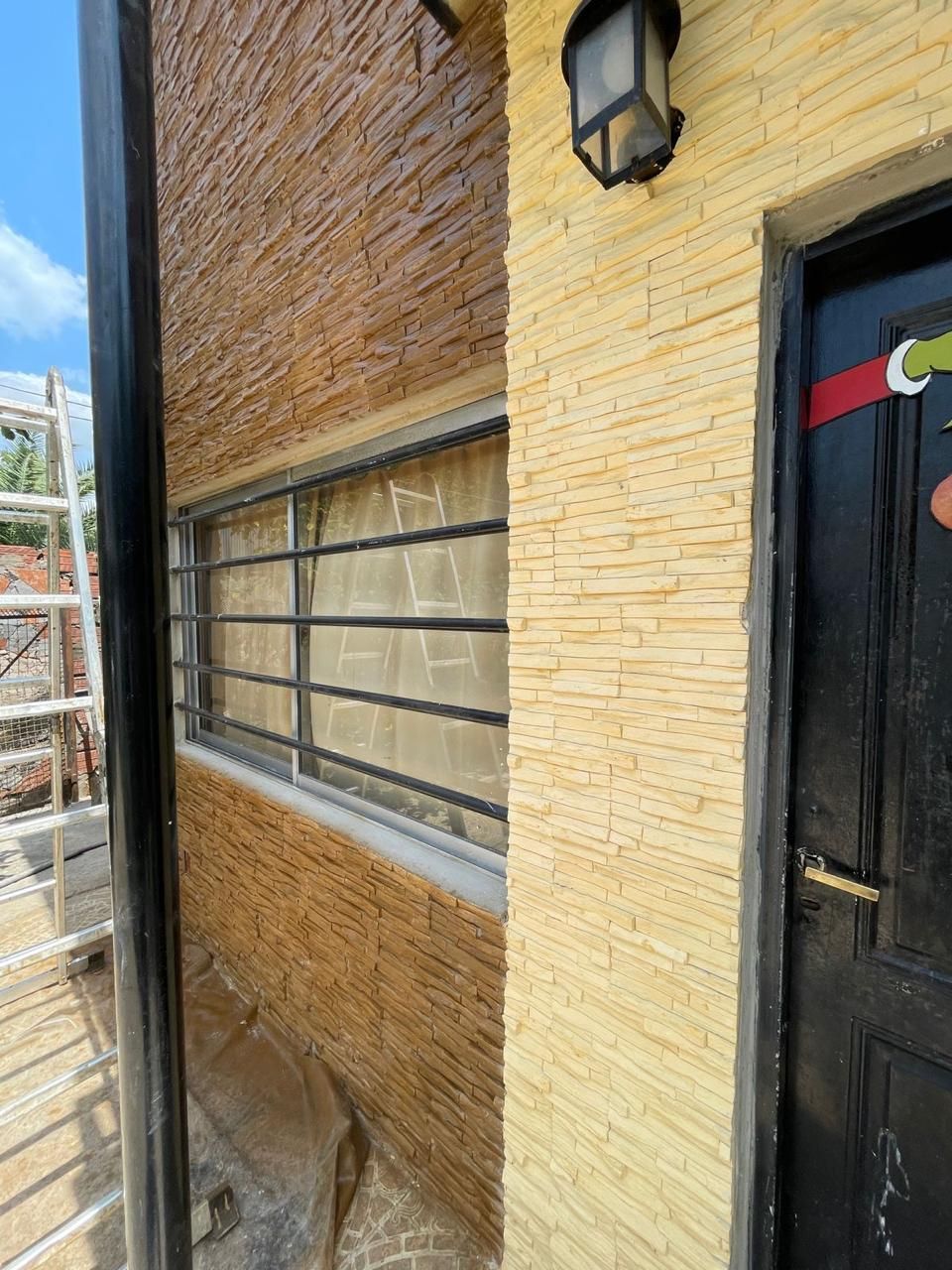 Exterior de un edificio con paredes texturizadas de color canela, una ventana con rejas, una puerta negra y una luz exterior.