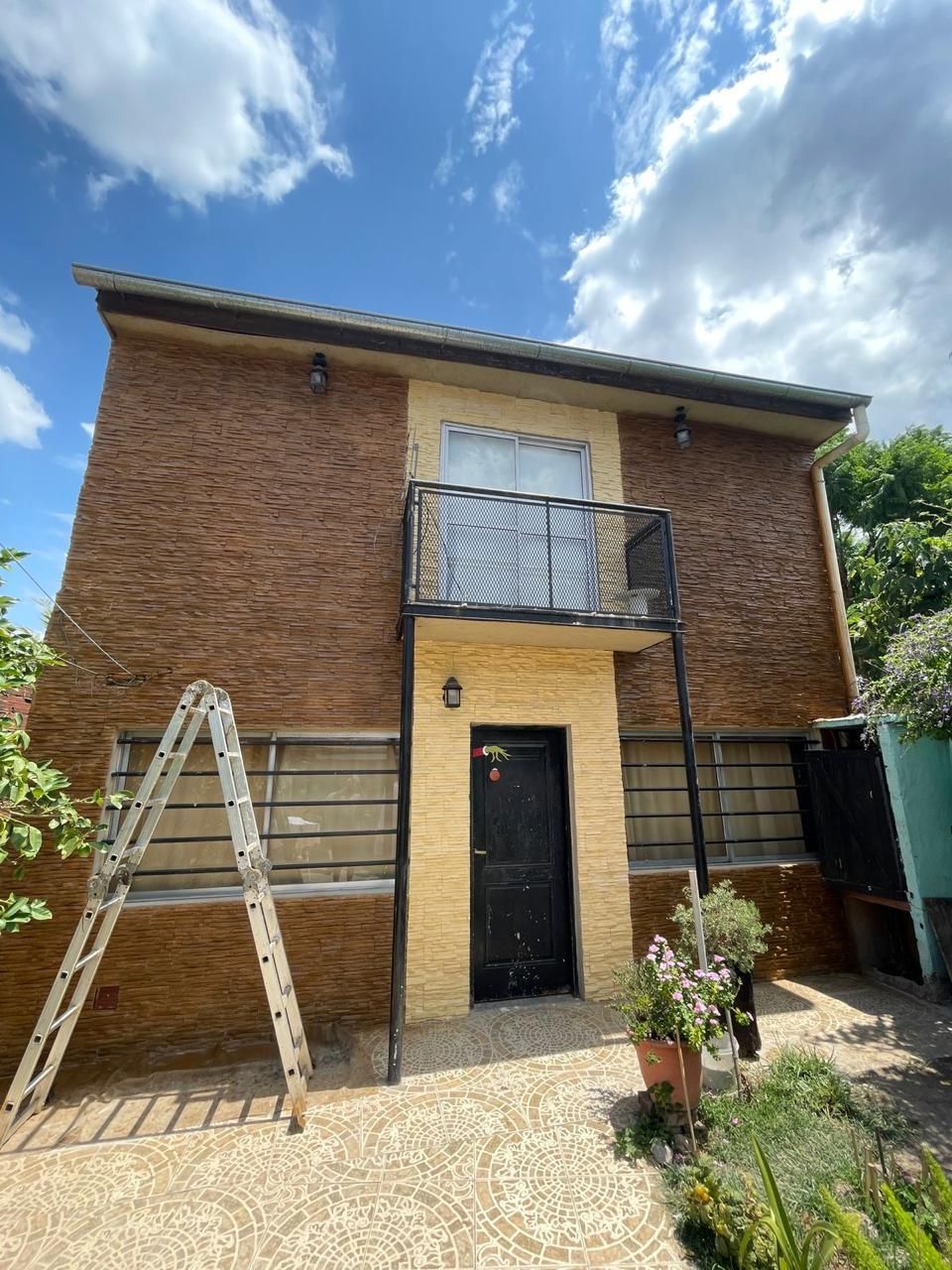 Edificio de ladrillo de dos plantas con balcón, puerta negra y escalera. Cielo azul con algunas nubes.