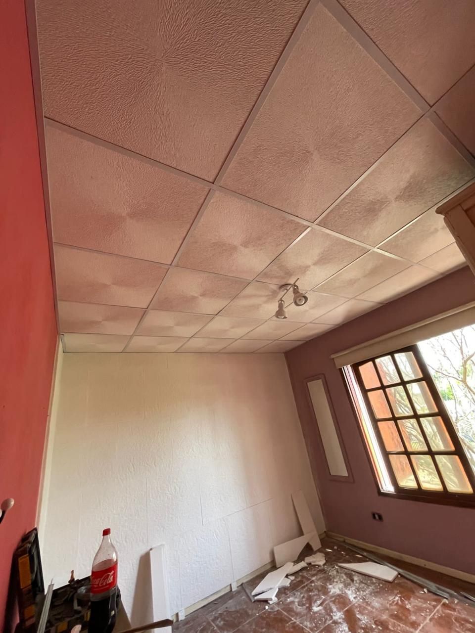 Una habitación con un techo texturizado y una ventana, las paredes son rojas y blancas, el piso está desordenado.