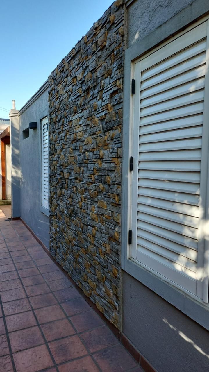 Sección de muro de piedra al lado de una pared gris con contraventanas blancas, sobre un patio de ladrillo.