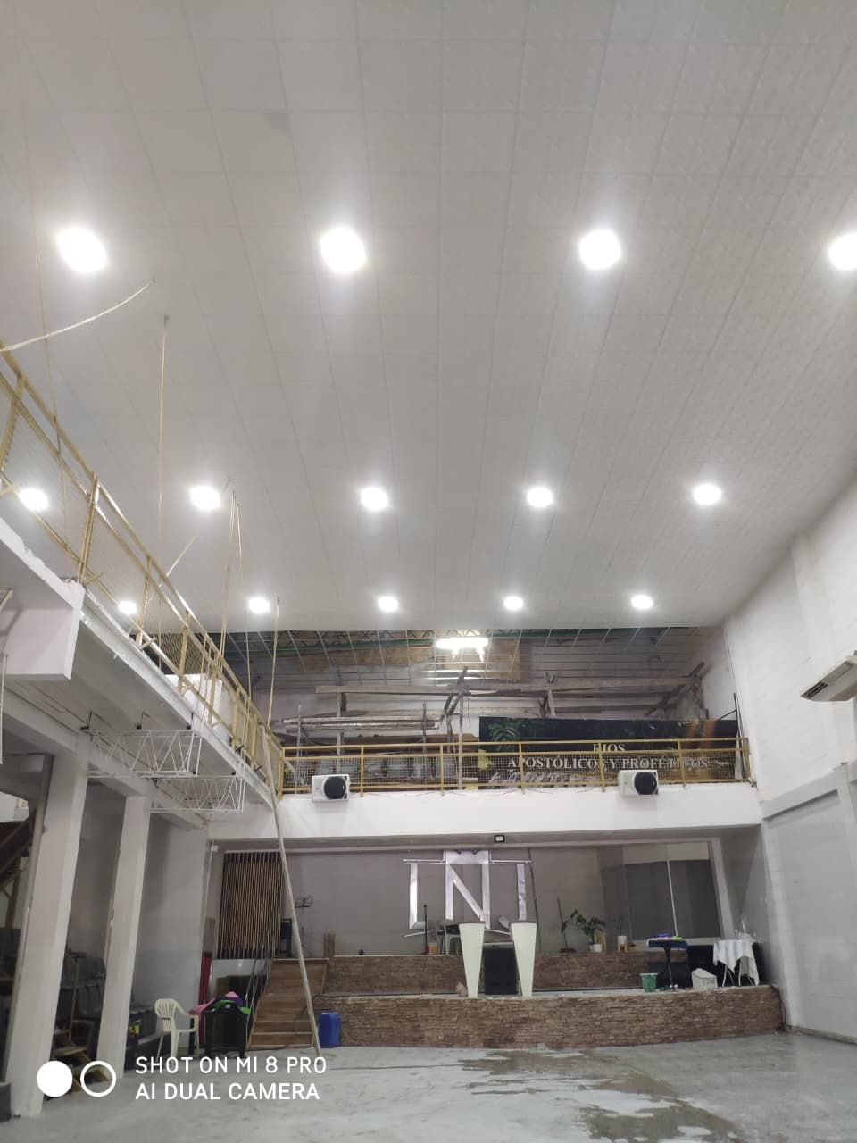 Interior espacioso y luminoso de un local con balcón, escenario e iluminación cenital. Paredes blancas y suelo de ladrillo.