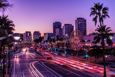 City street at dusk, long exposure of car lights, palm trees, Ferris wheel, and buildings under a purple sky.