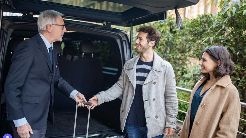 A chauffeur handing luggage to a smiling couple by a black van outdoors.