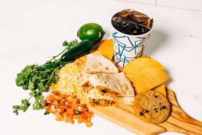 a wooden cutting board topped with quesadillas, chips, cookies, and a cup of soda
