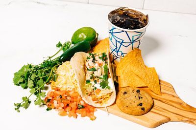 a wooden cutting board topped with a taco, chips, a cookie, and a cup of soda