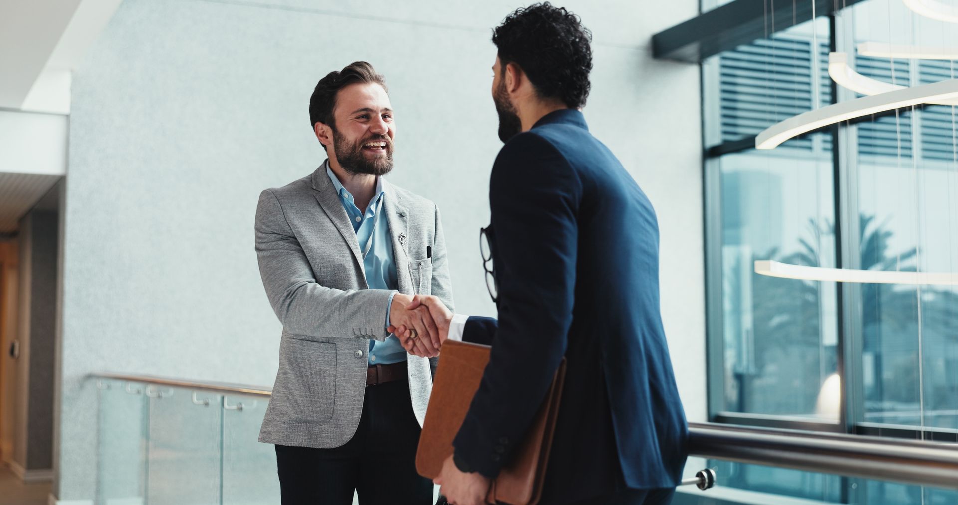 Two professionals shaking hands in a bright office hallway, one holding a folder.