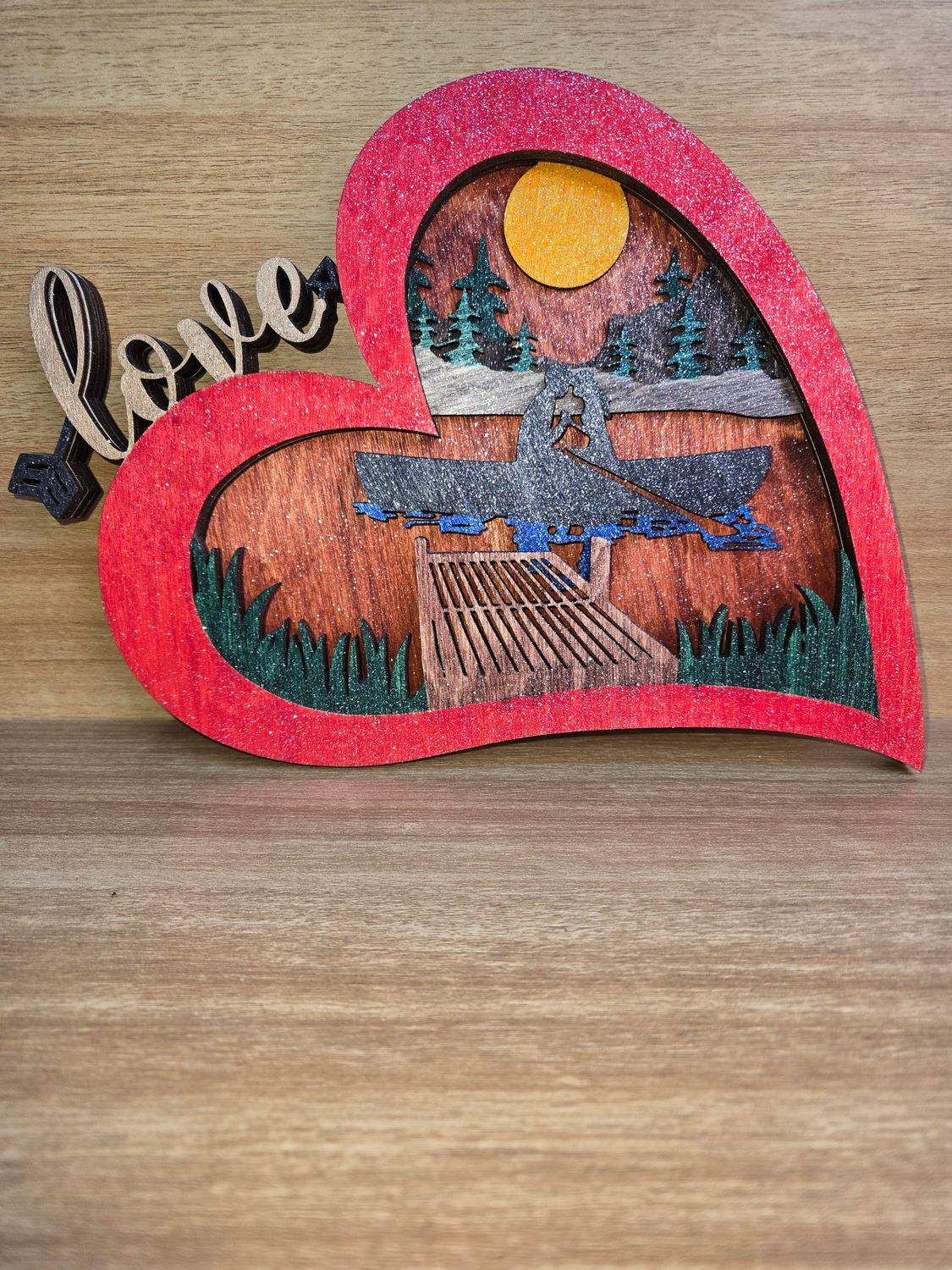 Red glitter heart with a wooden scene inside: dock, trees, sun, and the word 