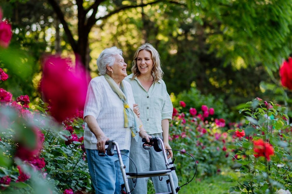 A Woman is Walking With an Elderly Woman With a Walker in a Garden — Eleos Support Services In Lavington, NSW