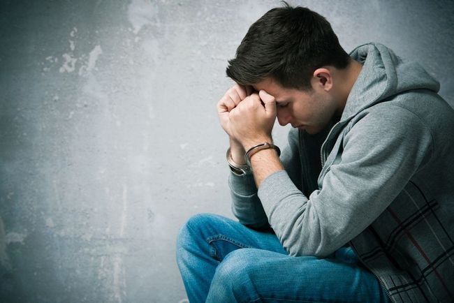 A young man sits with his head in his hands, appearing sad or distressed, in front of a textured gray wall.