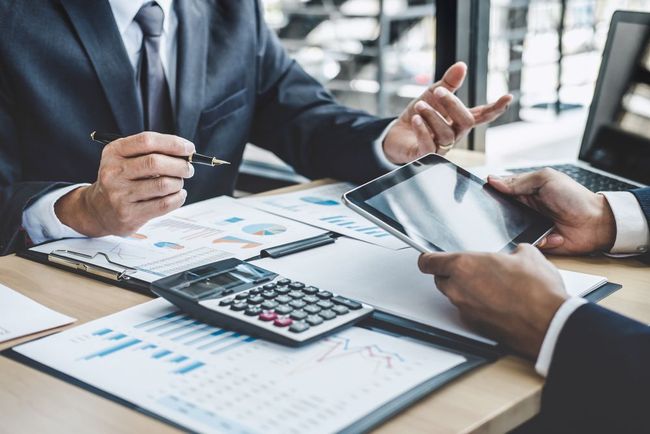 Two businesspeople in suits, discussing financial reports at a desk with a calculator and tablet.
