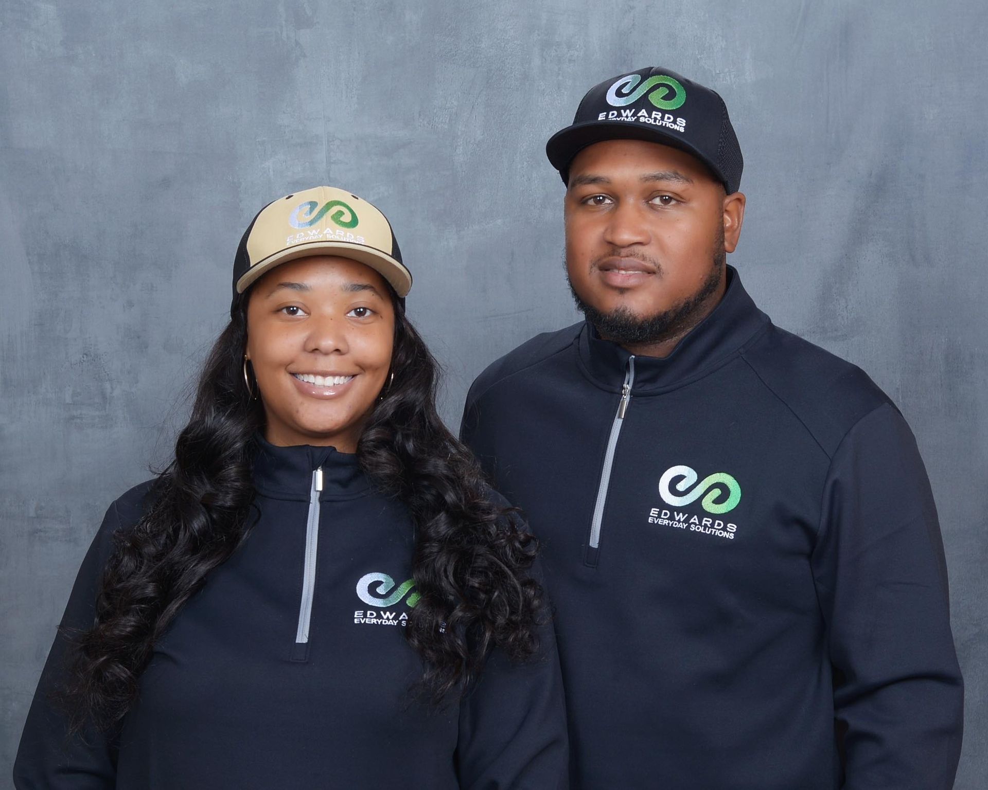 Two people posing against a grey background, wearing matching navy quarter-zip pullovers and branded baseball caps.