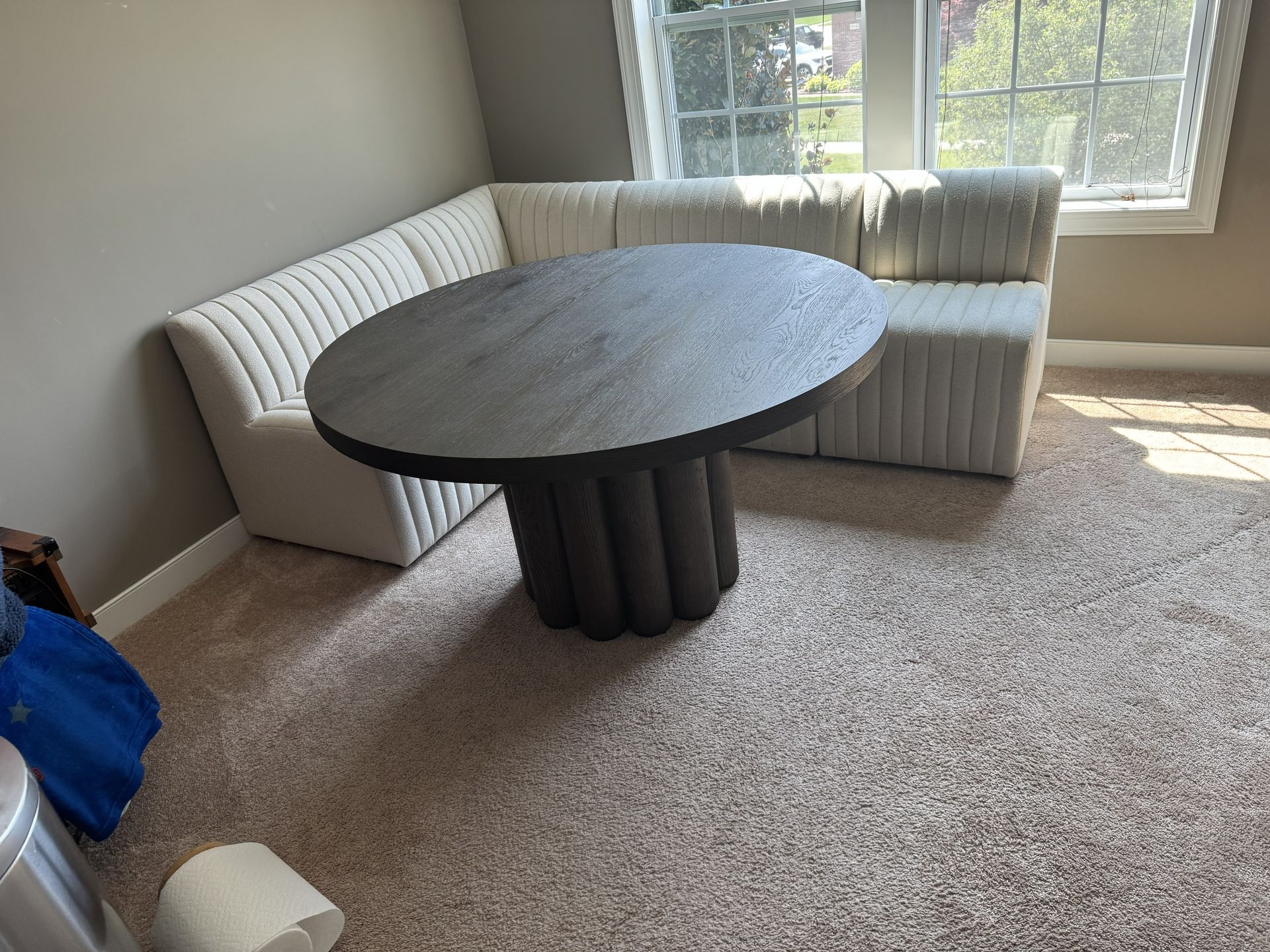 A dark circular pedestal table sits in front of a white upholstered corner booth in a carpeted room.