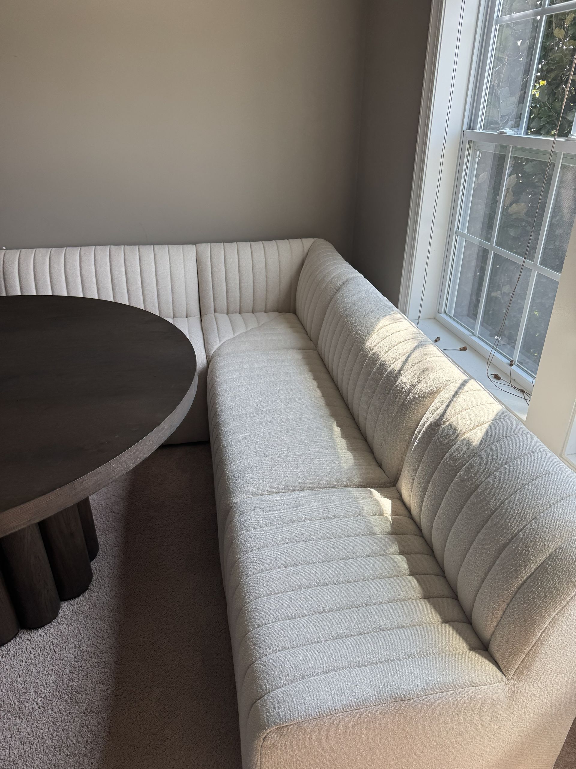A modern, light-colored, textured corner banquette sofa beside a circular dark wood pedestal table near a window.