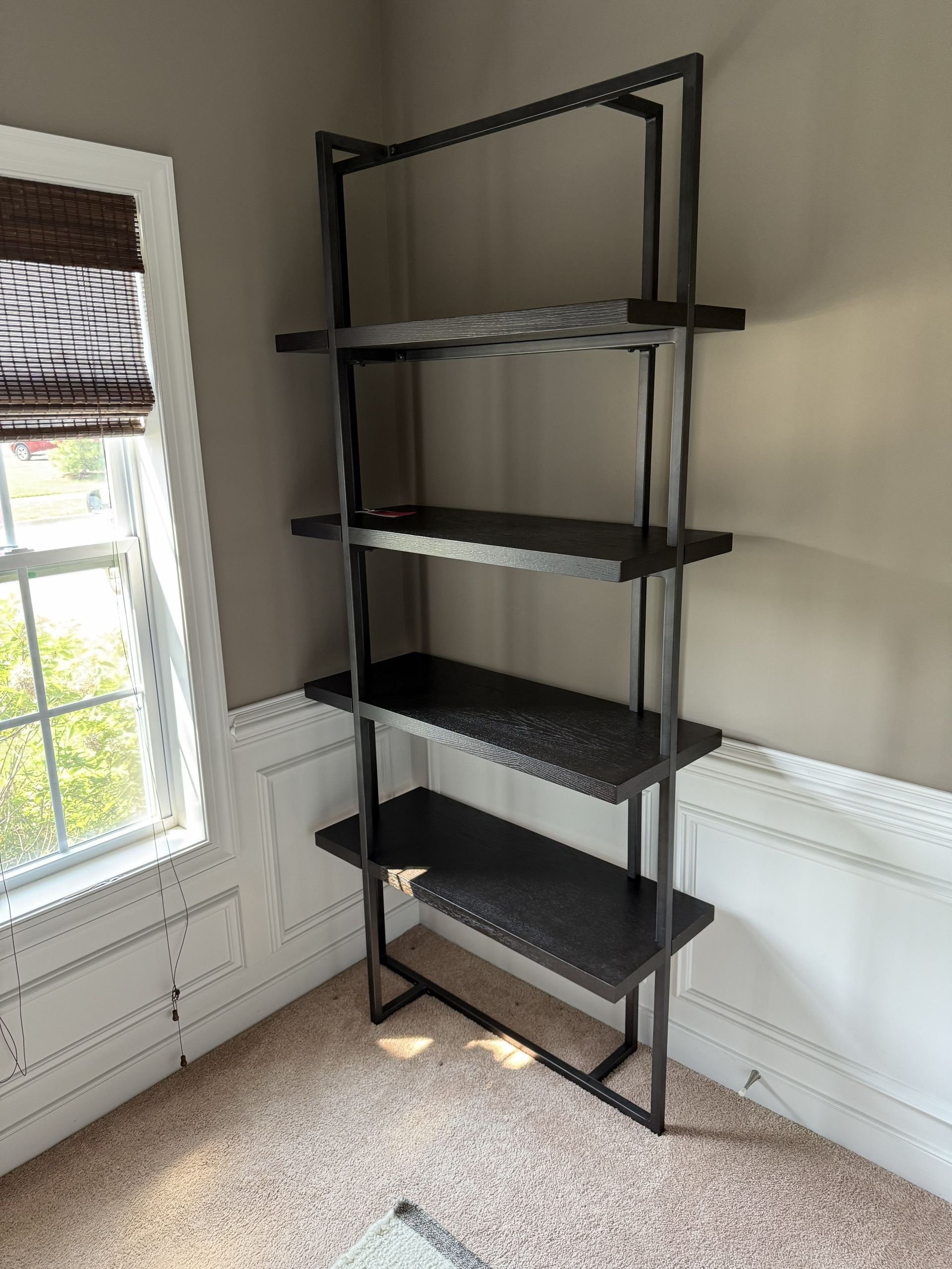 A tall, black industrial-style metal shelving unit with five shelves placed in the corner of a room with a window.