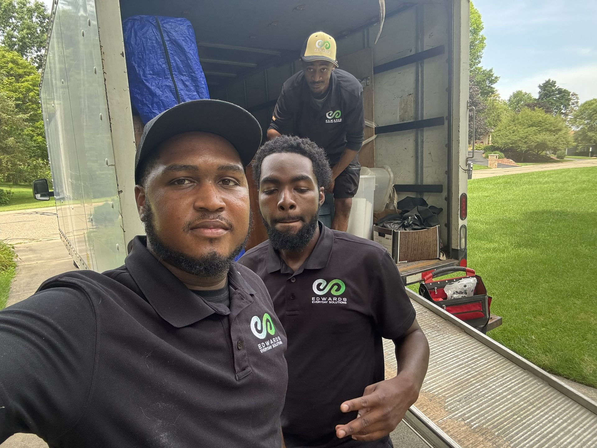Two movers in matching black company shirts take a selfie in front of an open moving truck in a grassy outdoor area.
